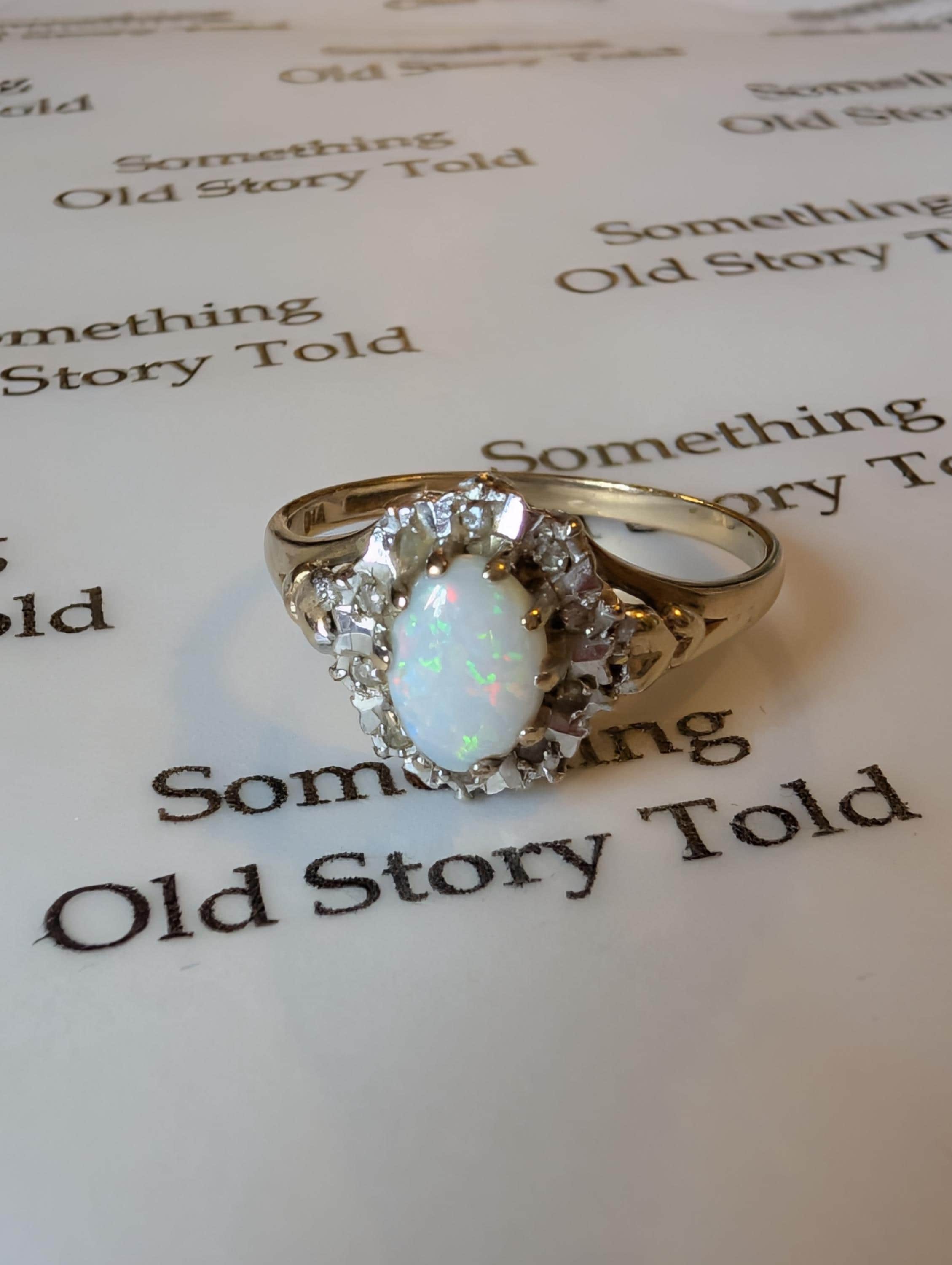 A gold ring with an oval opal gemstone and a halo of smaller diamonds The ring's band is polished gold. The background has the text "Something Old Story Told" printed on it. __etsy:7854008686