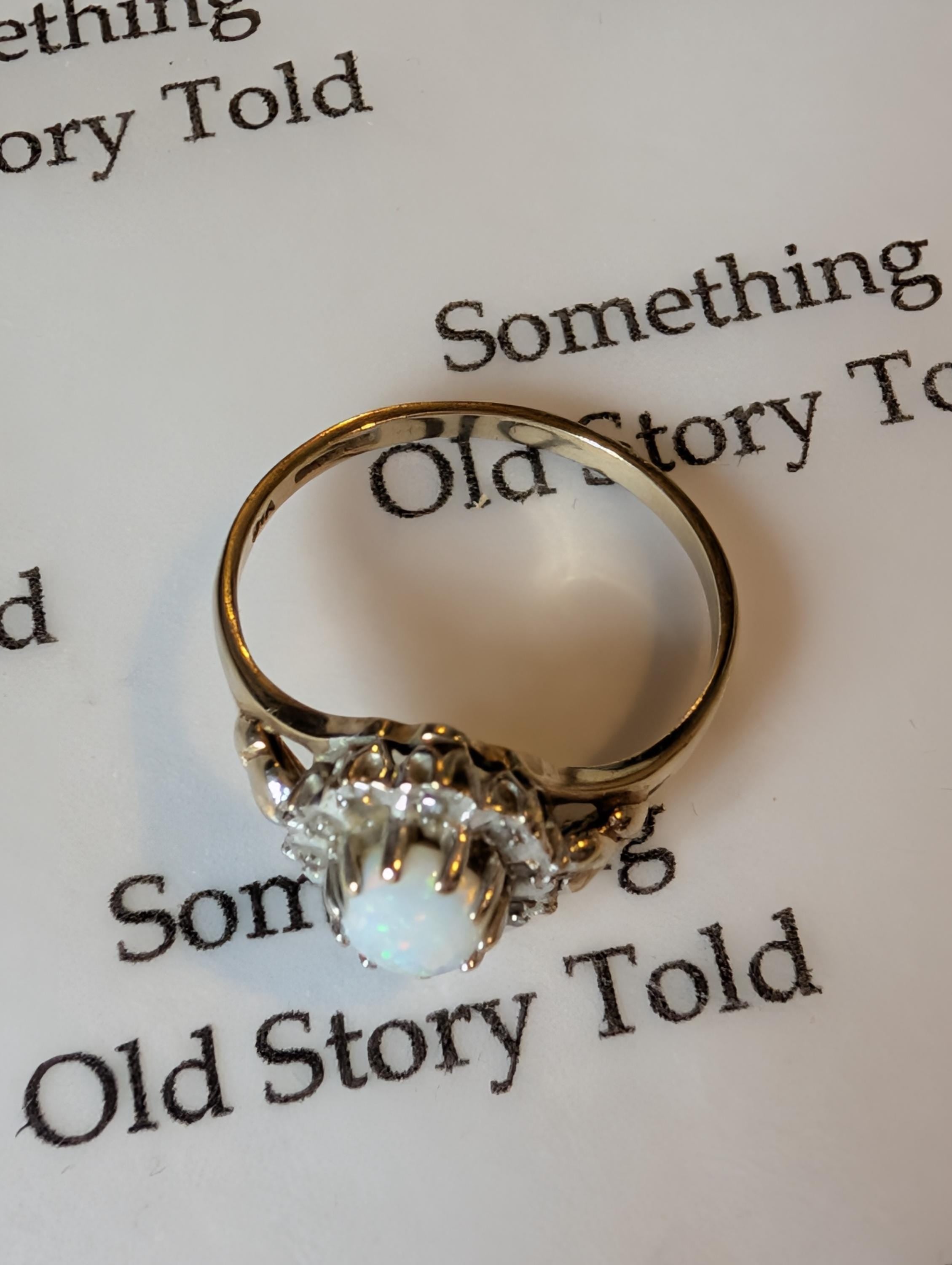 A gold ring featuring a central white opal gemstone, encircled by smaller diamonds. The ring rests on a white surface with the text "Something Old Story Told". __etsy:7901972015