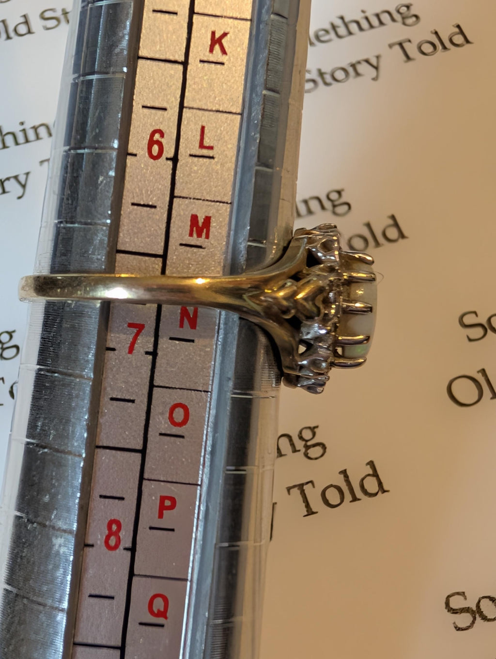 A gold ring featuring a large, oval, opalescent gemstone, encircled by smaller diamonds The ring is on a ring sizer, indicating a size between L . The ring has an ornate, vintage design. __etsy:7854010674