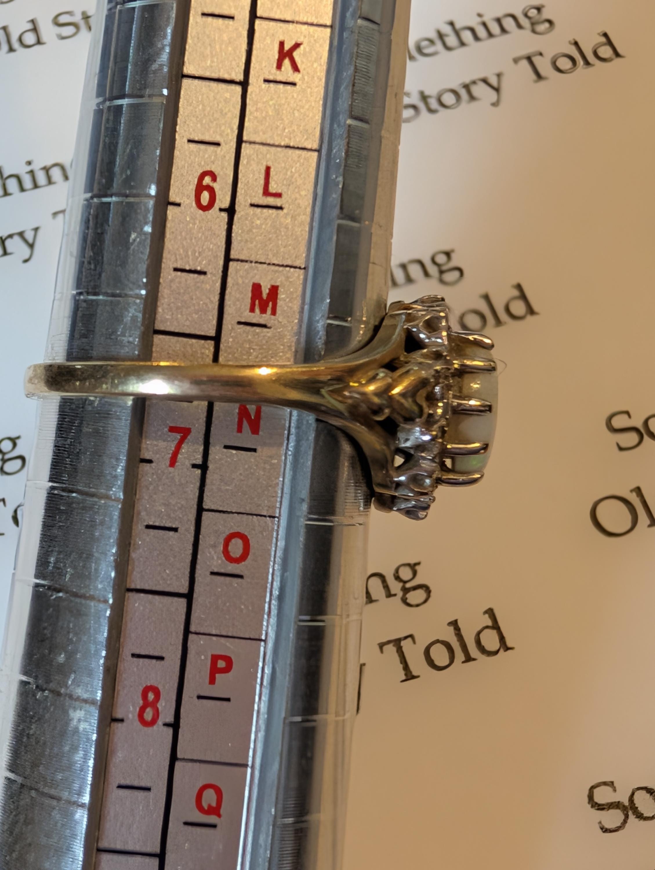 A gold ring featuring a large, oval, opalescent gemstone, encircled by smaller diamonds The ring is on a ring sizer, indicating a size between L . The ring has an ornate, vintage design. __etsy:7854010674