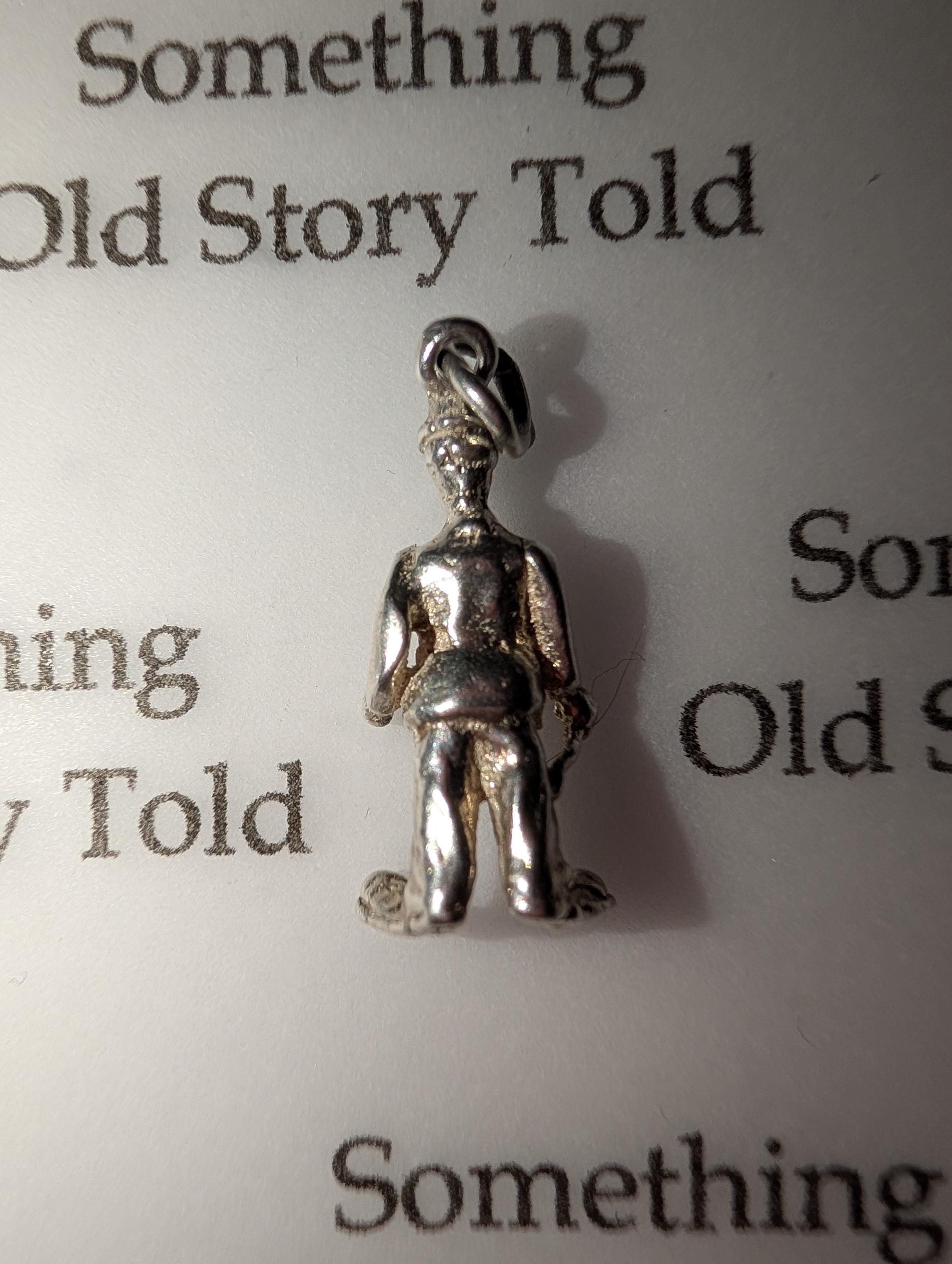 A small, silver-coloured charm depicting a person. The charm has a loop at the top for attachment and is standing on a white surface. The text "Something Old Story Told" is visible in the background. __etsy:7855648221