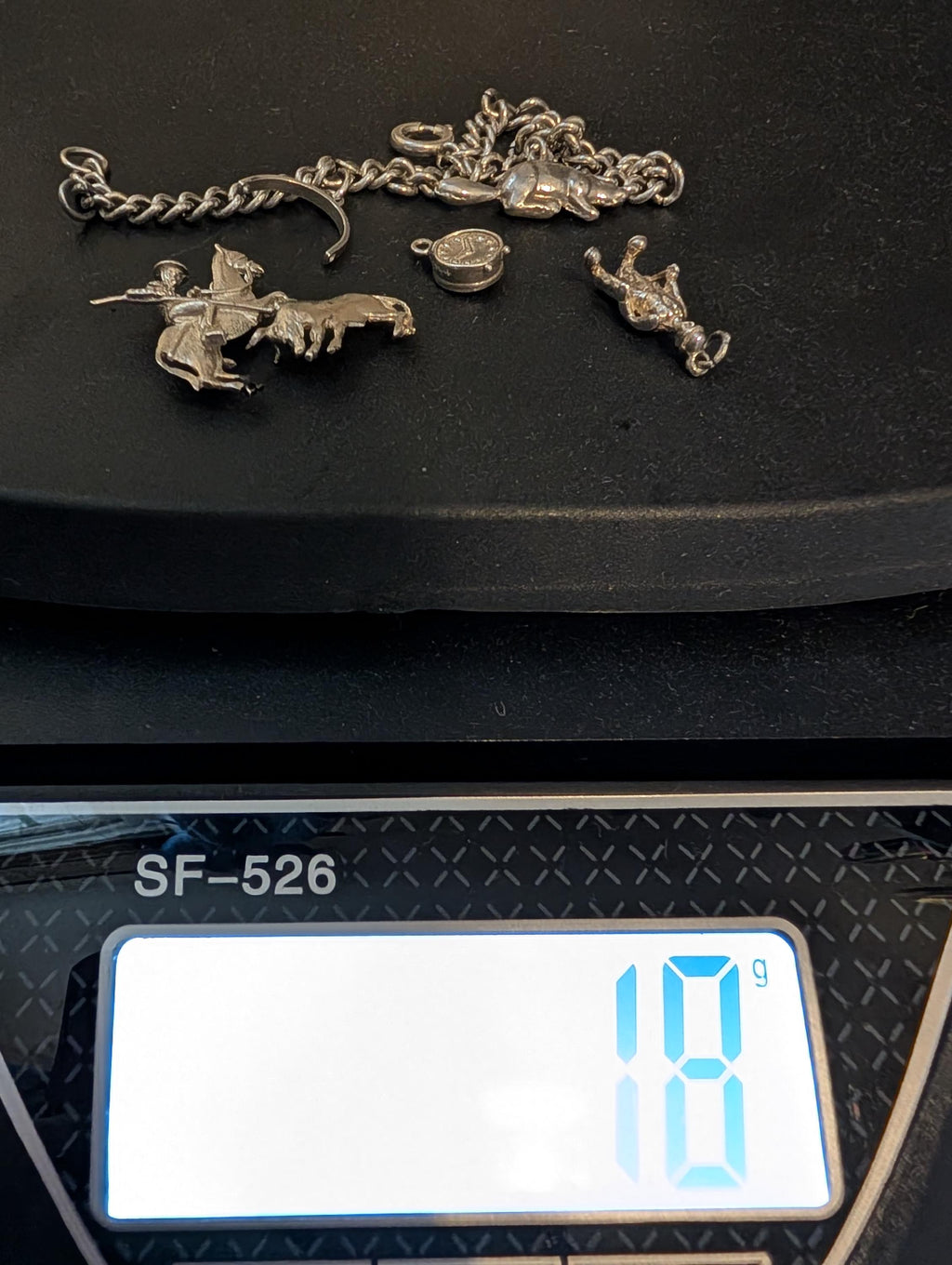 A selection of silver charms and a chain are presented on a black surface. The charms feature a rider on a horse, a bull, a crown, and a small round charm. The digital scale shows a weight of 18 grams. __etsy:7807707326