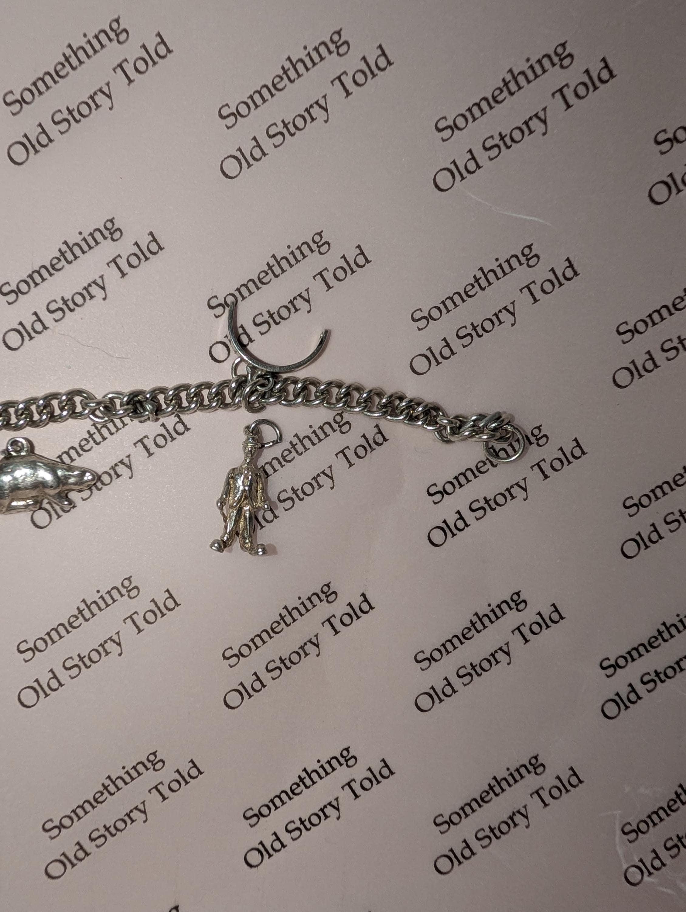 A silver charm bracelet with various charms. The bracelet has a chain link design and includes charms shaped like a small figure, a fish, and a circular clasp. The background has the repeated text "Something Old Story Told." __etsy:7807707310