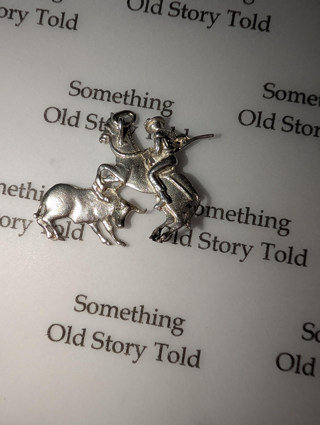 A silver charm showing a rodeo scene. It features a cowboy riding a bucking bronco, with a bull in the foreground. The charm is detailed and has a loop for attaching to a bracelet or necklace. The background has the text "Something Old Story Told". __etsy:7855648217