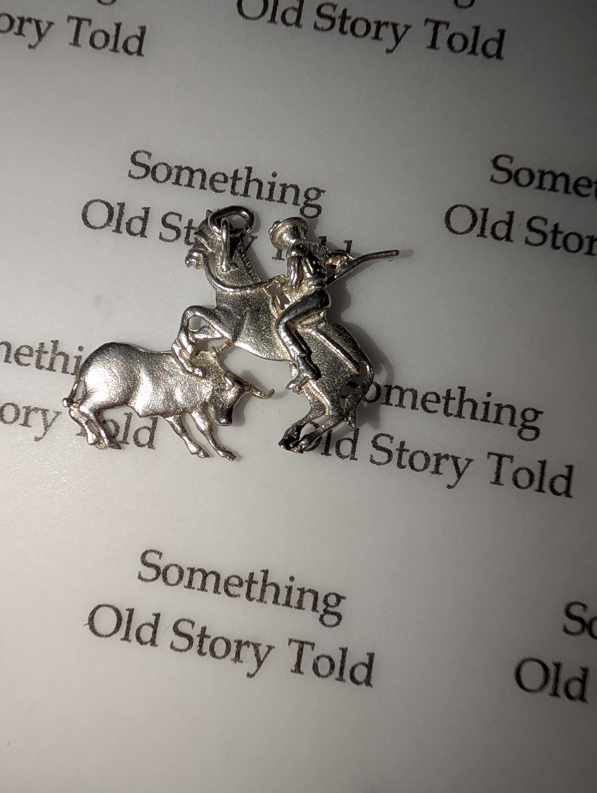 A silver charm showing a rodeo scene. It features a cowboy riding a bucking bronco, with a bull in the foreground. The charm is detailed and has a loop for attaching to a bracelet or necklace. The background has the text "Something Old Story Told". __etsy:7855648217