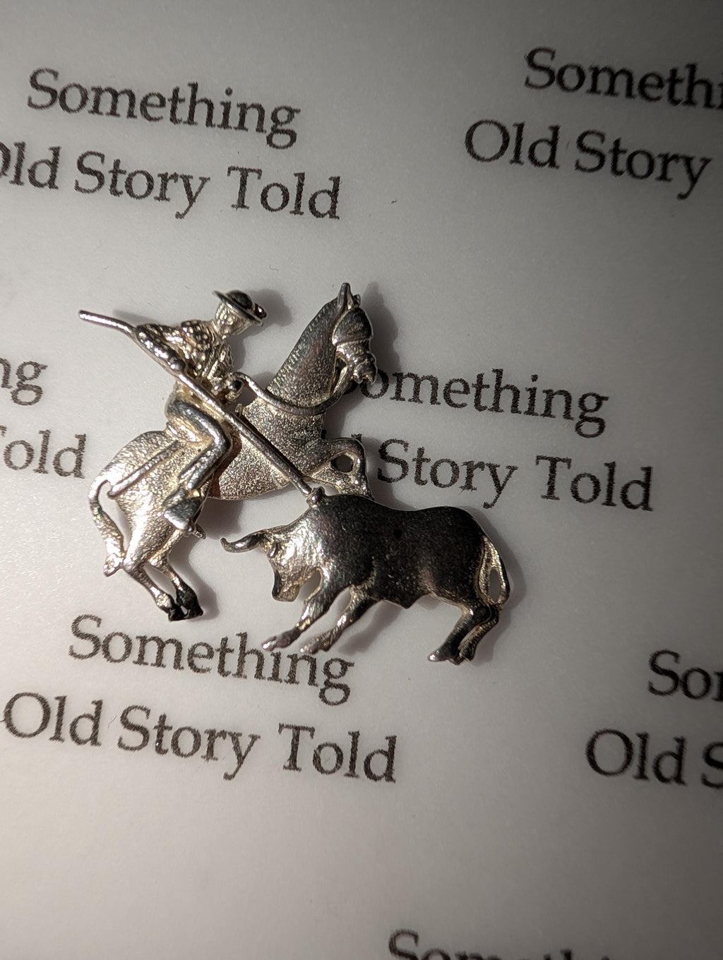 A silver charm featuring a matador on horseback and a bull. The charm has the text "Something Old Story Told" in the background. Ideal for jewellery making. The charm is made of silver-toned metal. __etsy:7807707318