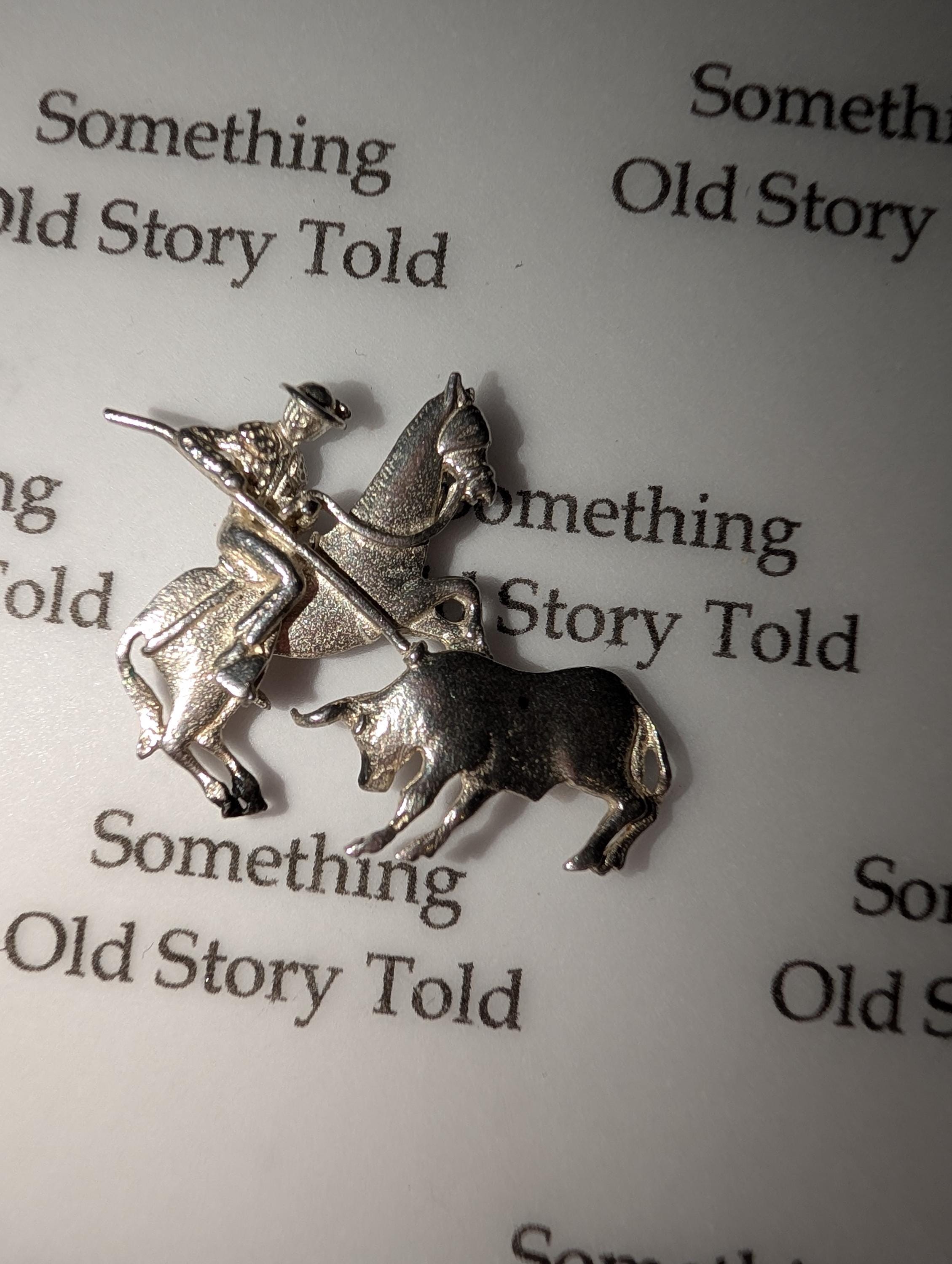 A silver charm featuring a matador on horseback and a bull. The charm has the text "Something Old Story Told" in the background. Ideal for jewellery making. The charm is made of silver-toned metal. __etsy:7807707318