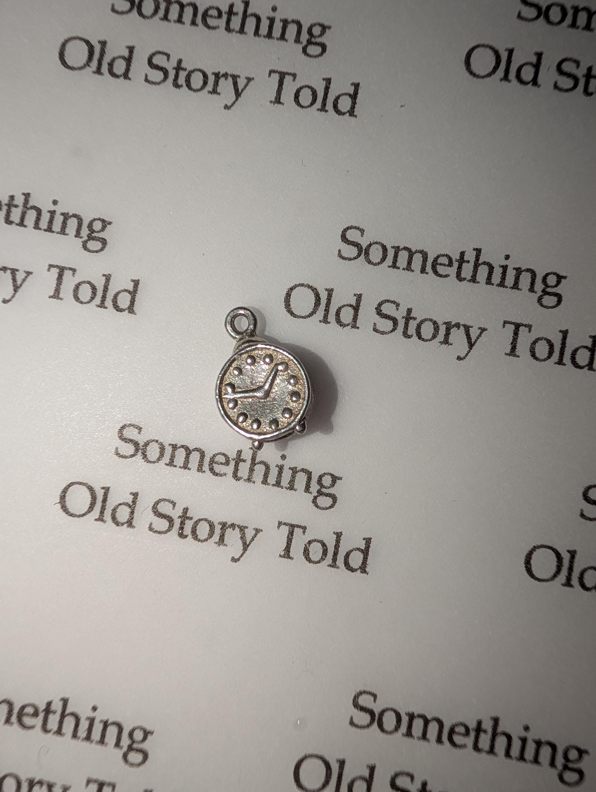 A small, silver clock charm with a loop for attaching to jewellery. The clock face has black markings and a dotted border. The words "Something Old Story Told" are printed on the background. __etsy:7855648175