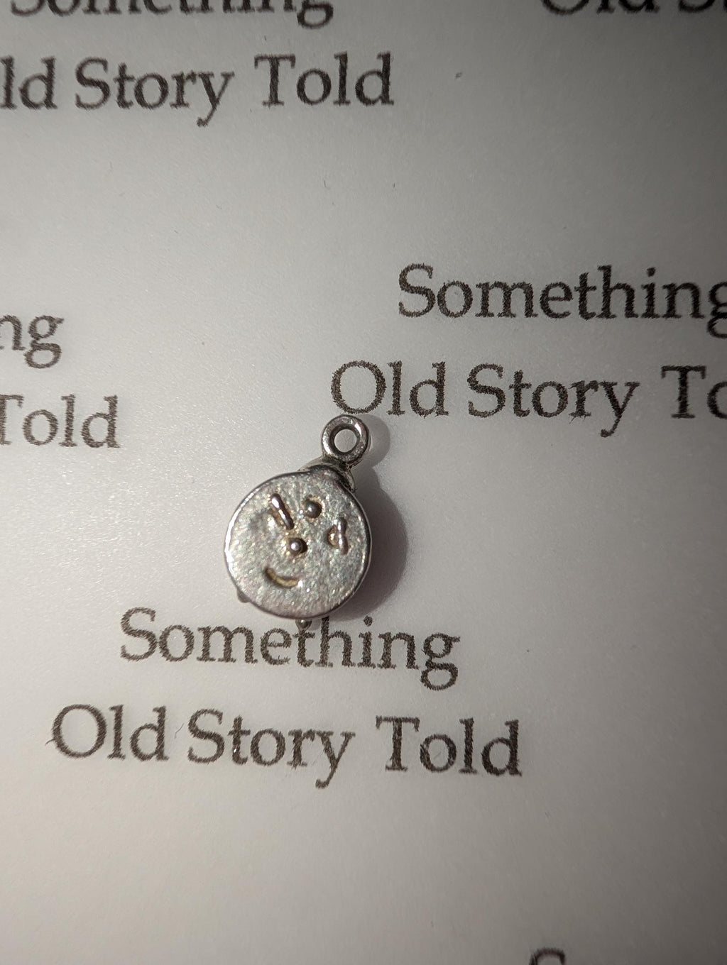 A small, silver charm with a smiley face design. The charm has a loop at the top for attaching to a necklace or bracelet. The text "Something Old Story Told" is printed on the background. __etsy:7855648205
