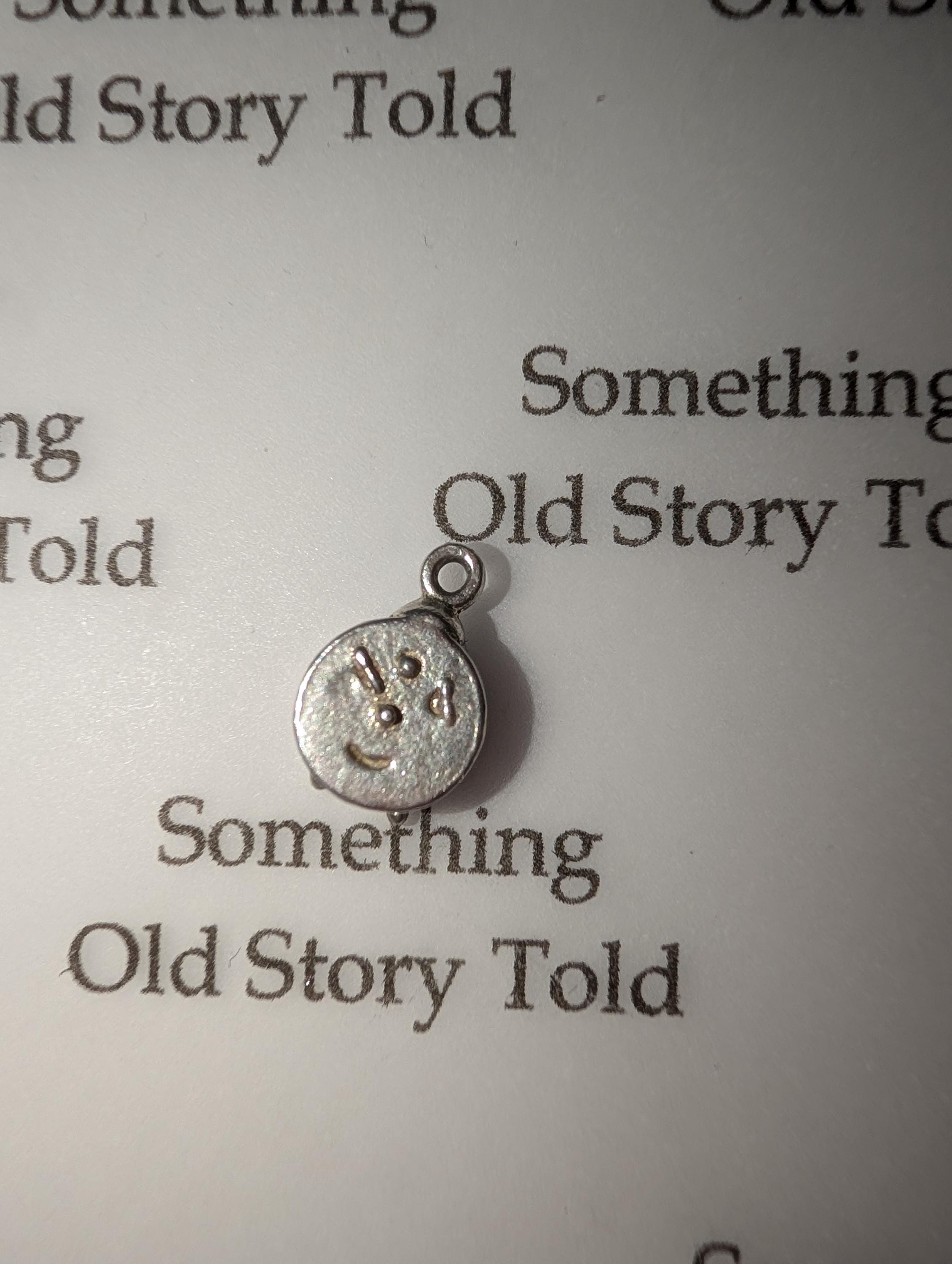 A small, silver charm with a smiley face design. The charm has a loop at the top for attaching to a necklace or bracelet. The text "Something Old Story Told" is printed on the background. __etsy:7855648205